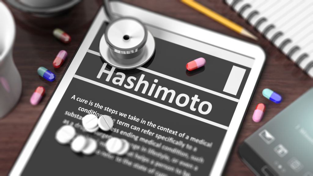 Tablet with "Hashimoto" on screen, stethoscope, pills and objects on wooden desktop.